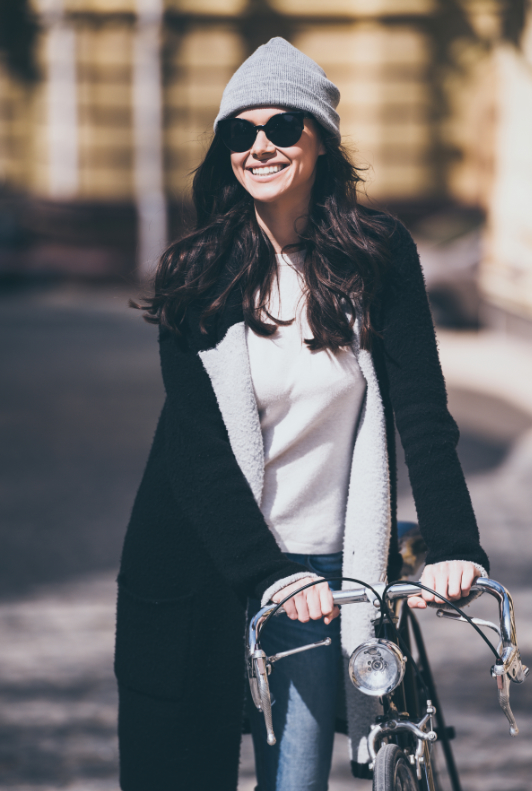 Kvinna gående genom stan med grå mössa, svart rock och en cykel