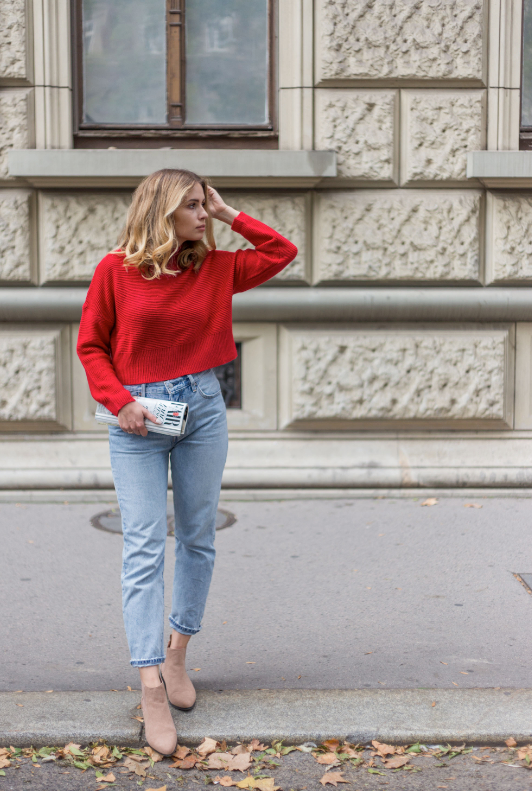 Vacker kvinna stående på gatan i en röd tröja och ljusa mom-jeans med ett magasin i handen