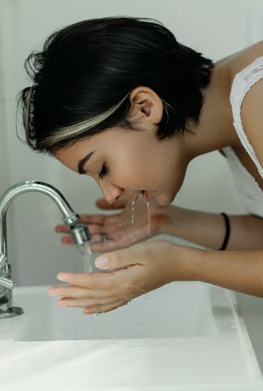 Svarthårig kvinna som står och tvättar ansiktet med vatten i handfatet