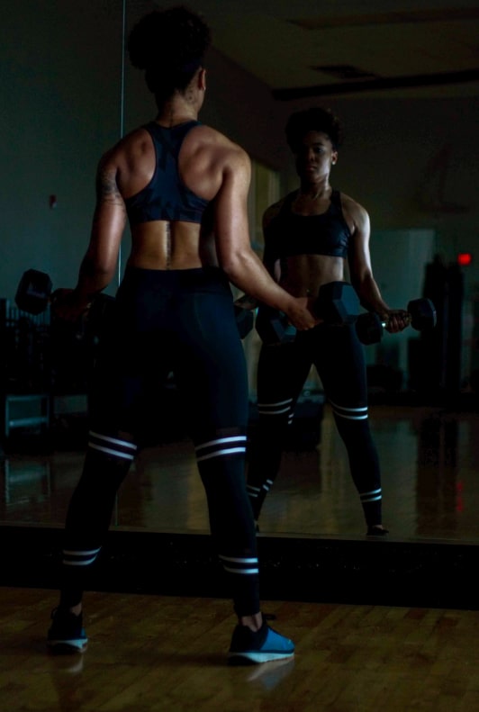 Kvinna som står framför spegelen med två hantlar i händerna i träningskläder
