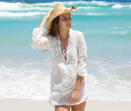Kvinna i vit strandklänning stående på strand med turkost hav i bakgrunden